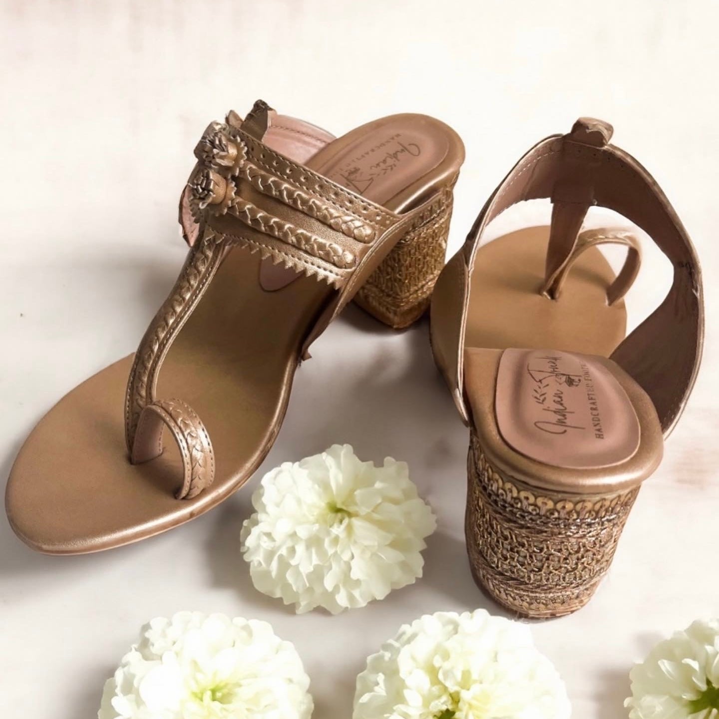 Pair of gold sandals with intricate designs on a white background with flowers.