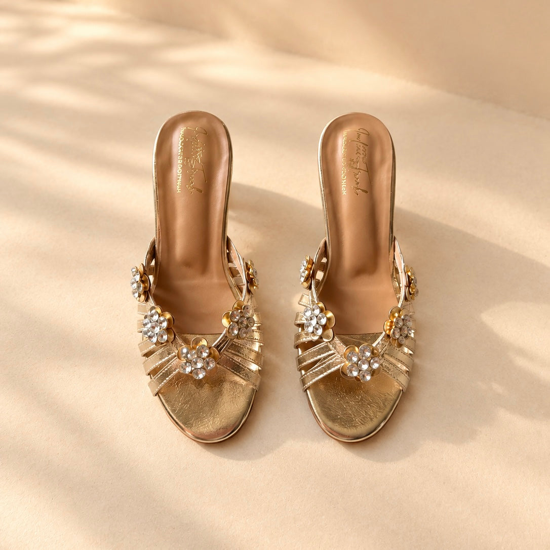 A pair of metallic gold heeled sandals with jeweled floral embellishments on the straps, displayed on a beige surface with soft leaf shadows.