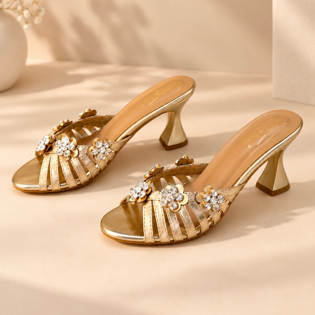 A pair of metallic gold heeled mule sandals with floral embellishments and rhinestone details, displayed on a warm beige surface with soft shadows.