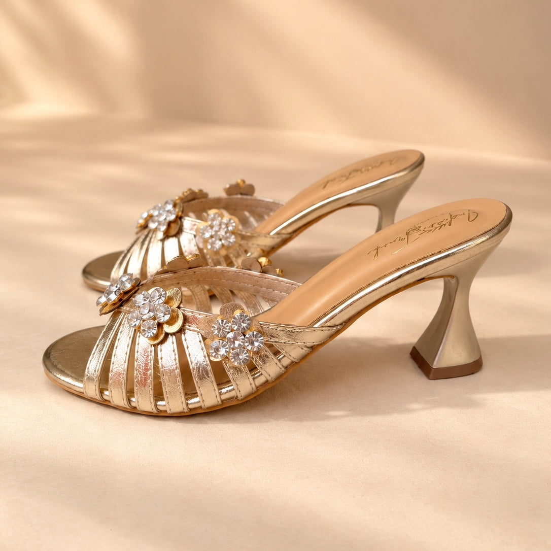 A pair of metallic gold heeled mule sandals with cutout straps and crystal-embellished floral details, displayed on a beige fabric background.