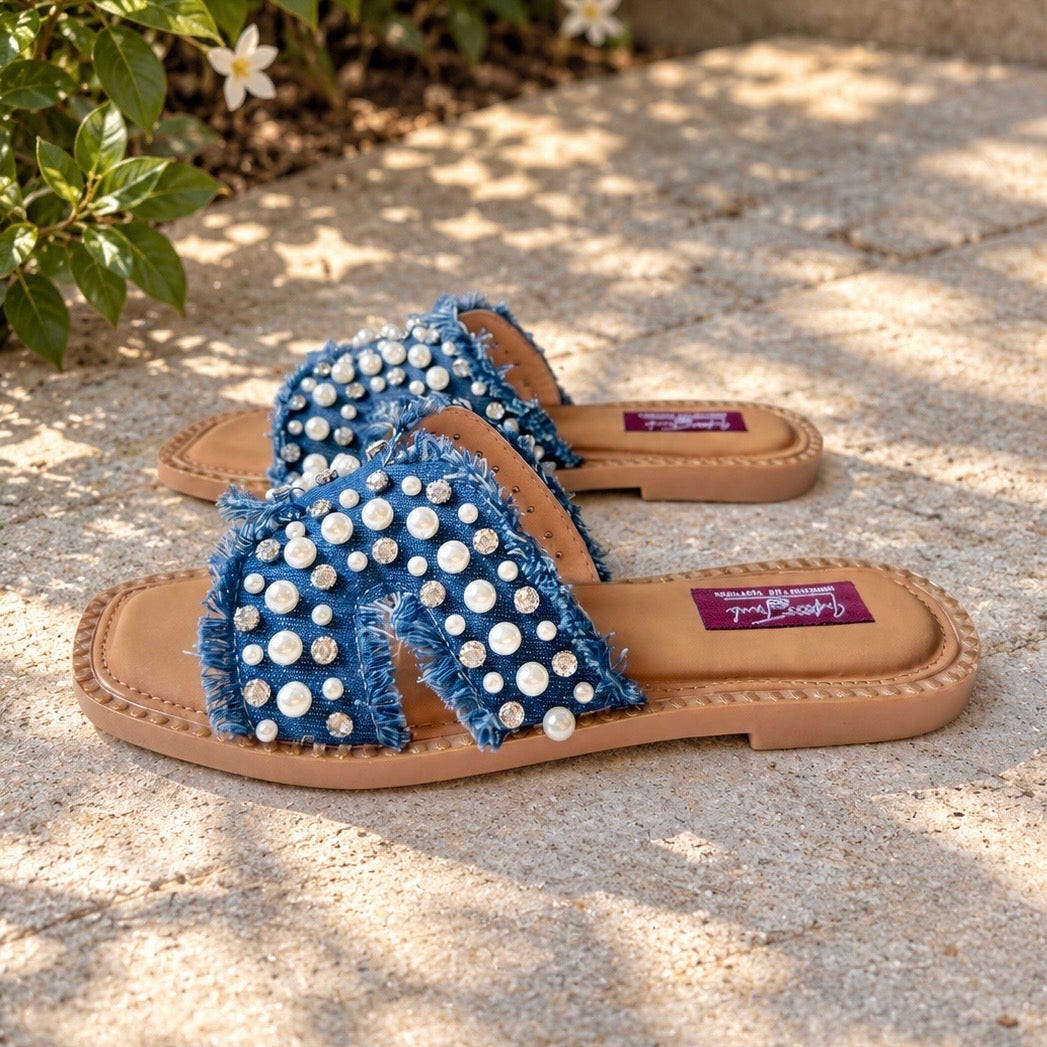 A pair of women's slide sandals with frayed blue denim straps embellished with white rhinestones, resting on a sunlit stone patio.