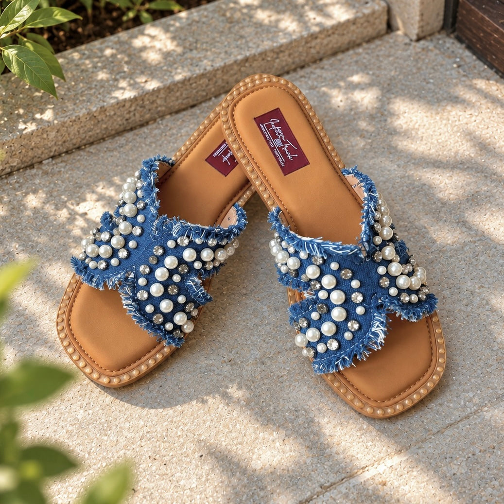 A pair of blue denim slide sandals embellished with white faux pearls and rhinestones, resting on a light gray stone patio with natural dappled sunlight and green leaves in the foreground.