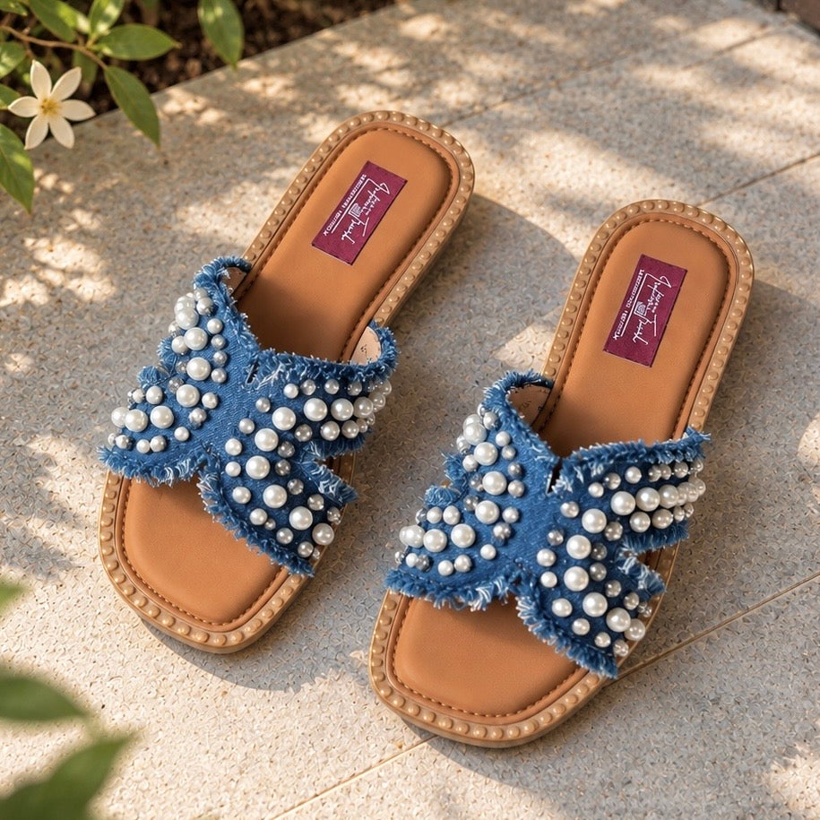 A pair of blue frayed-leather slide sandals embellished with white faux pearls, resting on a light stone patio with dappled sunlight and green leaves in the foreground.