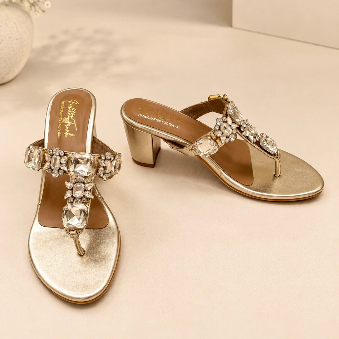 A pair of metallic gold block-heel thong sandals decorated with sparkling clear rhinestones and crystals, displayed on a light beige surface.