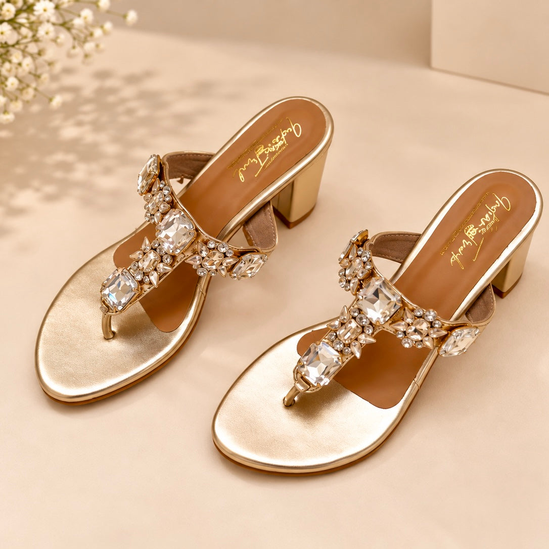 A pair of metallic gold heeled mule sandals with crystal-embellished toe straps, displayed on a beige surface with a soft floral prop in the corner.