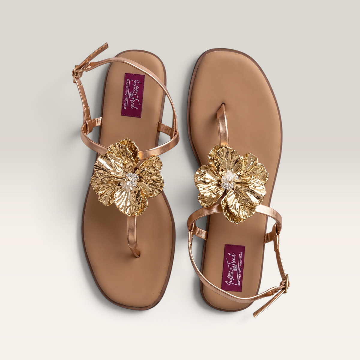 A pair of metallic rose gold flat sandals with large gold flower embellishments on the toes, photographed from a top-down angle on a light gray background.