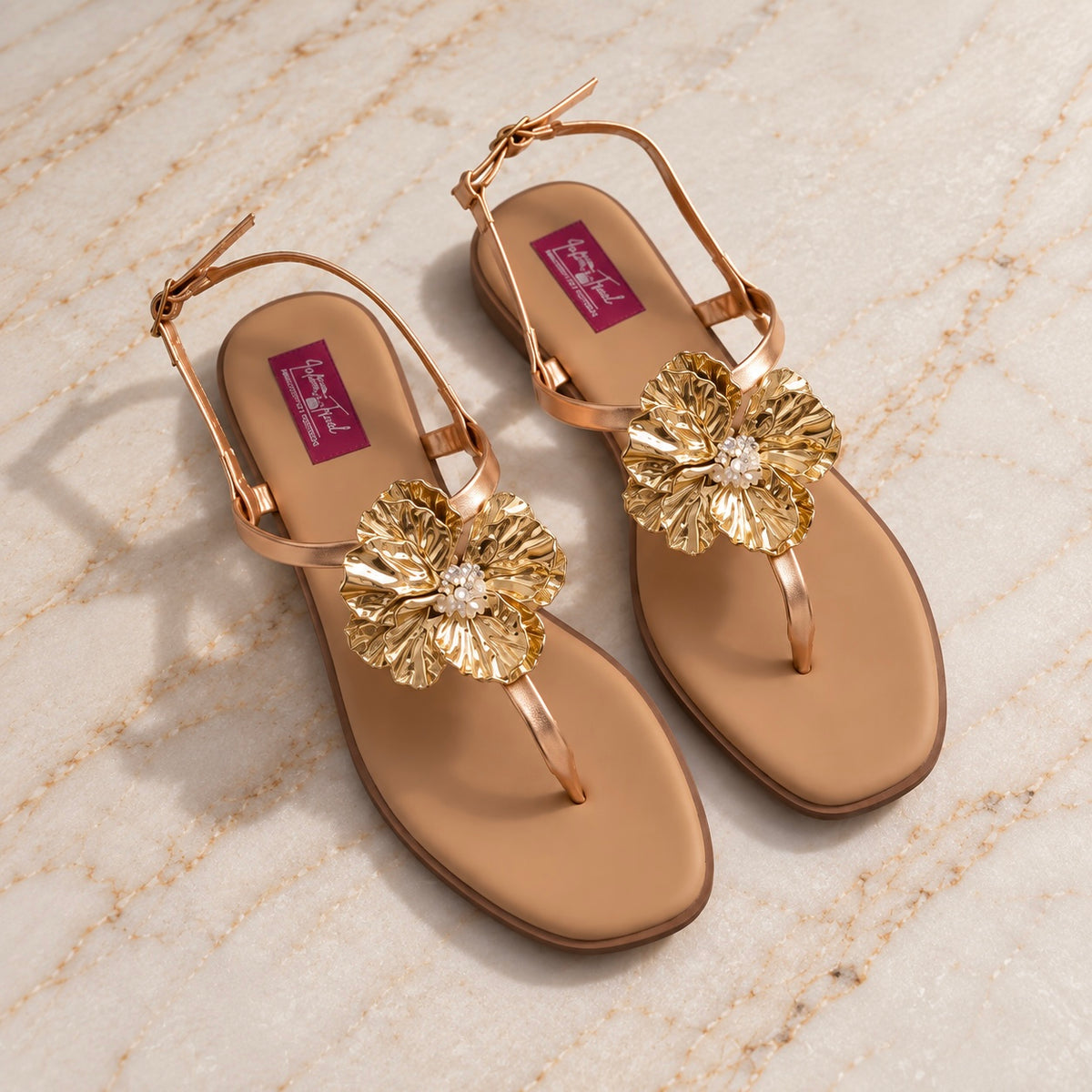 A pair of gold-toned flat sandals with metallic flower embellishments and rhinestone centers, displayed on a light beige marble surface. The insoles feature a magenta brand label with white lettering.