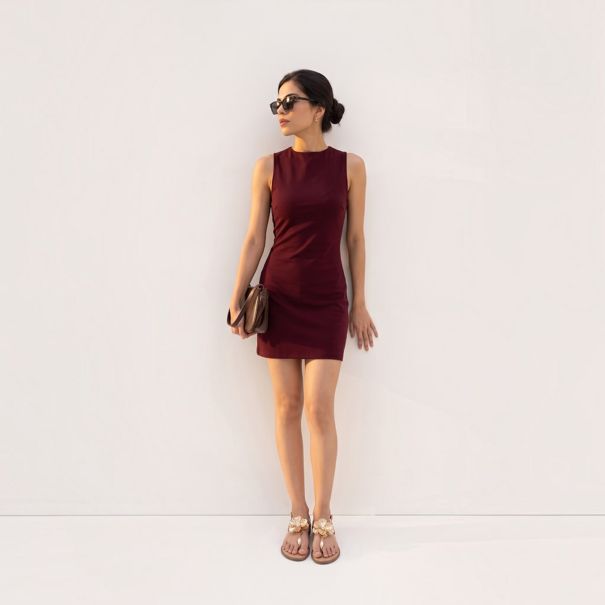 A woman stands against a plain white wall wearing a sleeveless, bodycon mini dress in deep burgundy. She is also wearing sunglasses, a small brown clutch, and flat sandals.