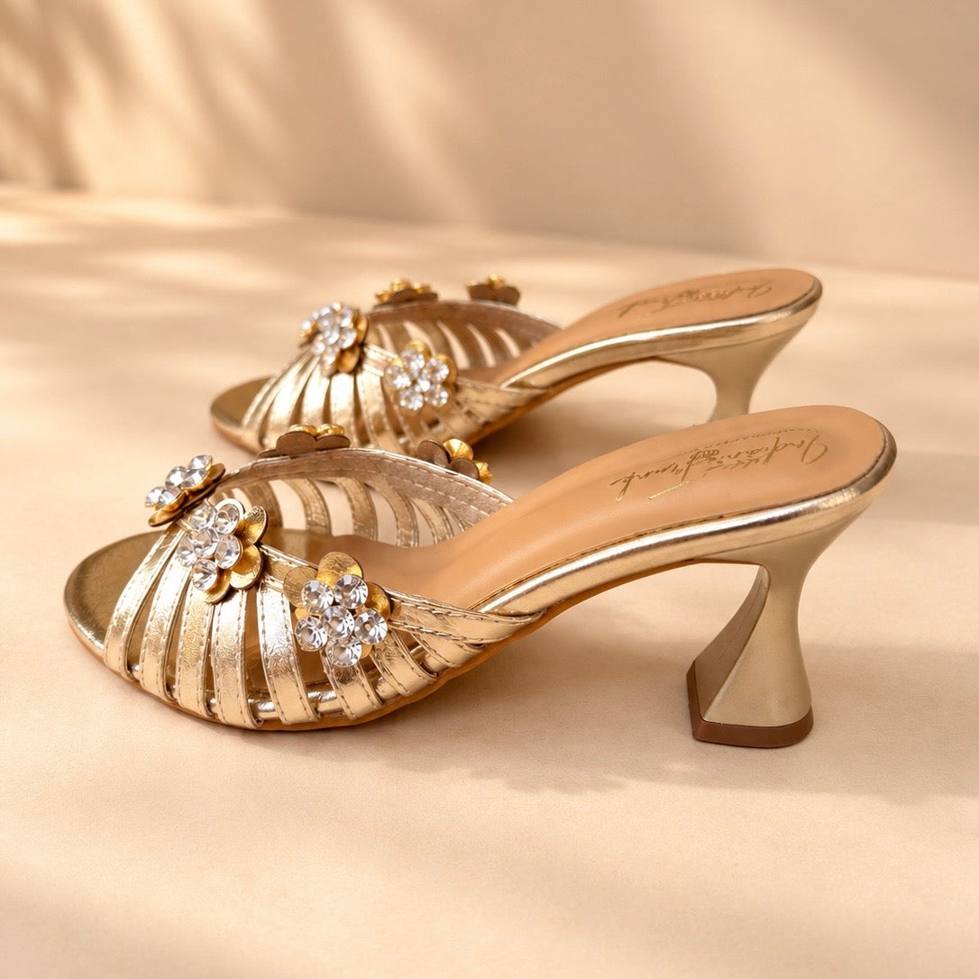A pair of metallic gold heeled mule sandals with floral embellishments and rhinestone accents, displayed on a beige fabric background.