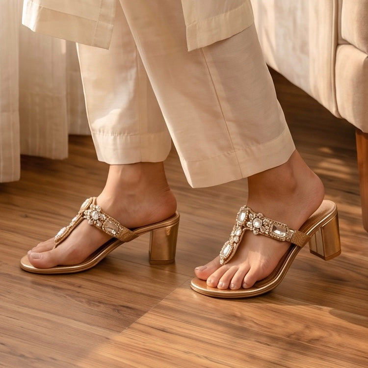 A pair of women's block-heel thong sandals in metallic gold with ornate crystal embellishments on the straps, shown being worn on a person's feet on a wooden floor.
